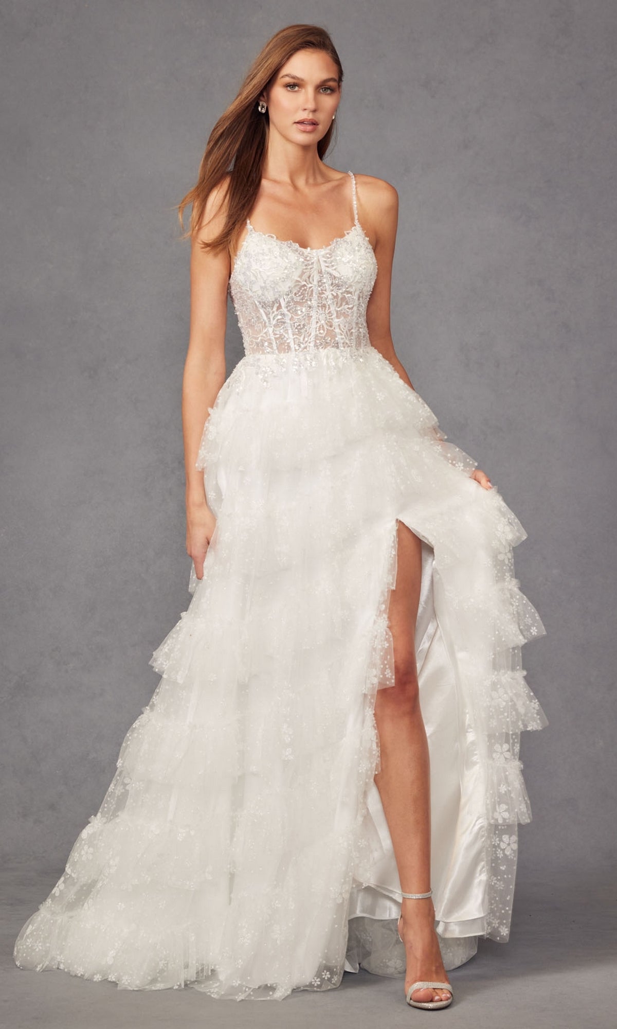 Woman wearing the Long Prom Dress JT2464AW by Juliet—an off-white lace gown with spaghetti straps, delicate tiers, and a high slit—posing against a gray background for an elegant, timeless look.